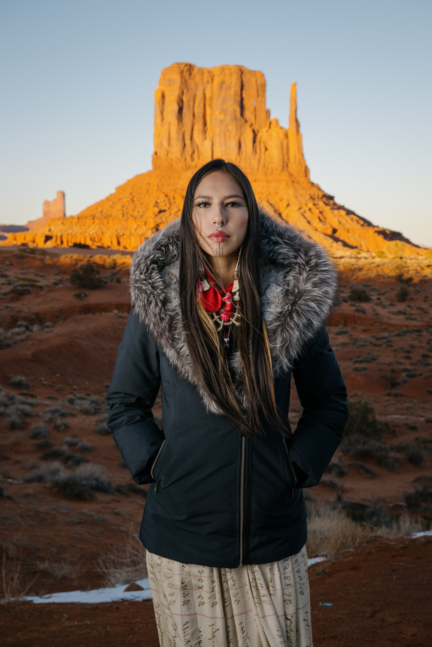Portrait of Quannah Rose Chasinghorse-Potts, in front of sacred Indigenous land near her childhood home on the Navajo Nation. The landform is known as West Mitten, in Monument Valley, Utah. Set aside in 1958, it is a Navajo Nation tribal park, considered sacred by Diné and managed by the nation. Self-management of lands sacred to Indigenous peoples is one major aspect of the Native American landback movement.